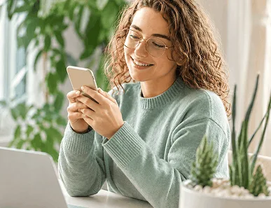 Vrouw met bril kijkt naar smartphone aan tafel met laptop en planten